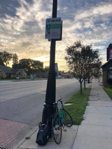 An image of a DDOT bus stop with a bike leaning against it. It is morning and the clouds are luminous with sunlight
