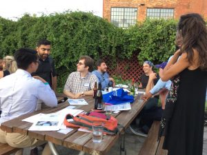 A group of people talking in several smaller pairings around a picnic table on a patio. Some people sit and some stand. There are grapevines on the fence in the background.