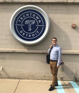 A man in a blue shirt and khakis stands smiling on a sunny sidewalk next to the circular TechTown Detroit logo posted on the wall.