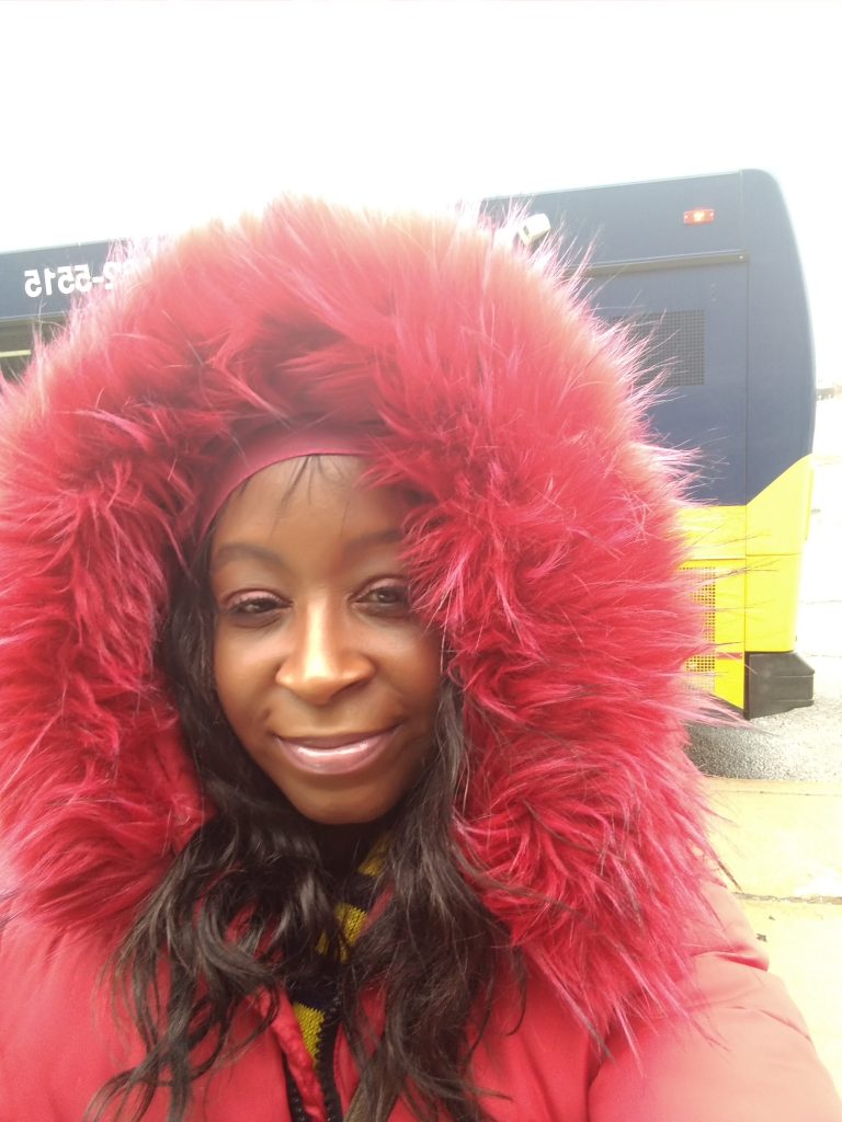 LaShondra standing in front of a SMART bus, wearing a jacket with a bright red fur hood, with her hair looking amazing