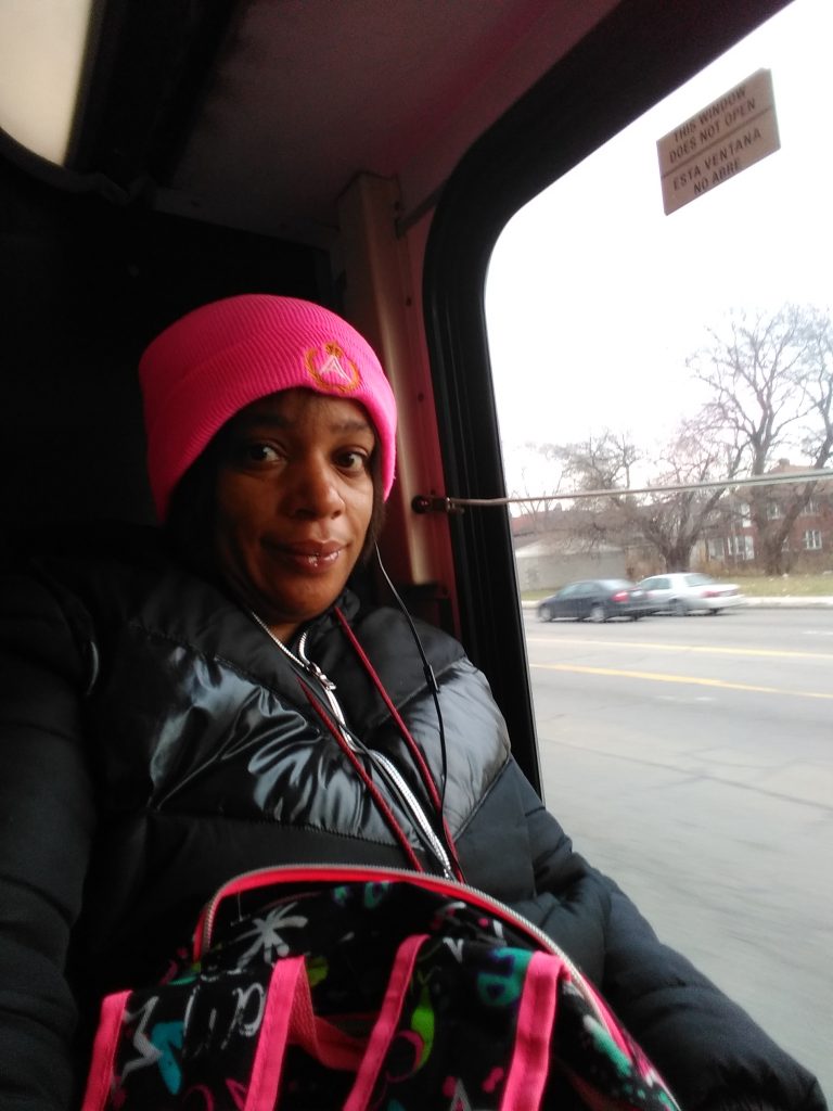 Tonika Williams sitting on a bus next to the window. She is looking at the camera. Her hat and the trim of her backpack are the exact same shade of hot pink.