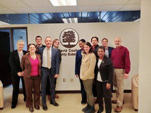 Twelve people stand in front of the Oakland County Executive crest, including County Executive Dave Coulter and TRU's Megan Owens