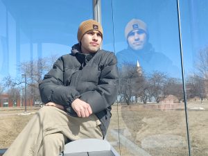 Thomas Yazbeck sits on a bench in a bus stop, reflected by the glass. He is wearing a yellow hat and a gray coat. The sky is brightly blue and he looks into the distance.