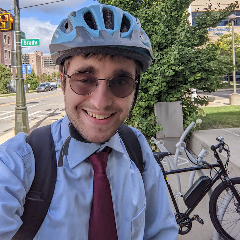 Corey in a shirt and tie wearing a helmet with his bike in the background