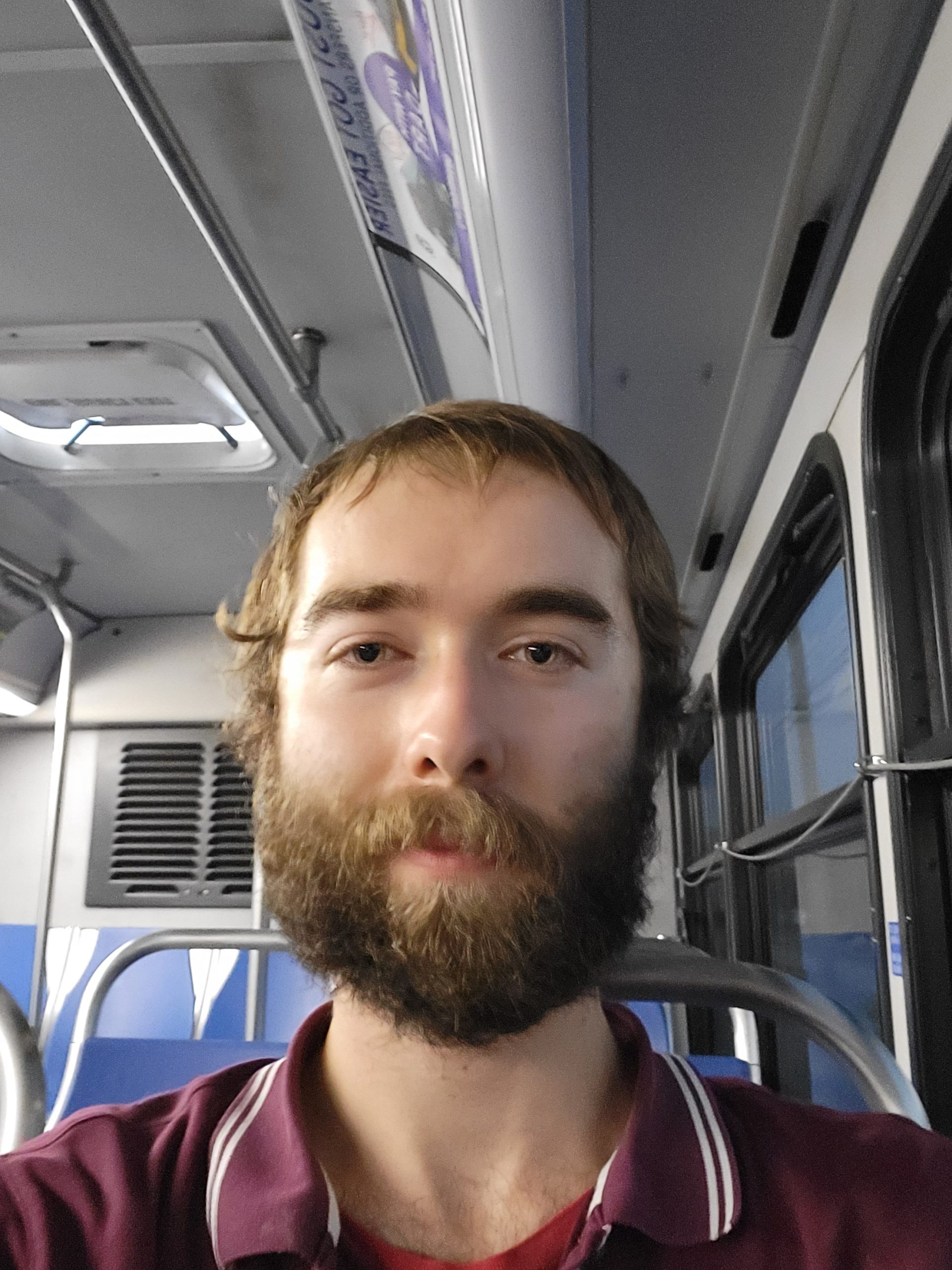 Photo of Alex Selsey, a white man with dirty blonde hair and a full beard sitting on a bus in a red polo