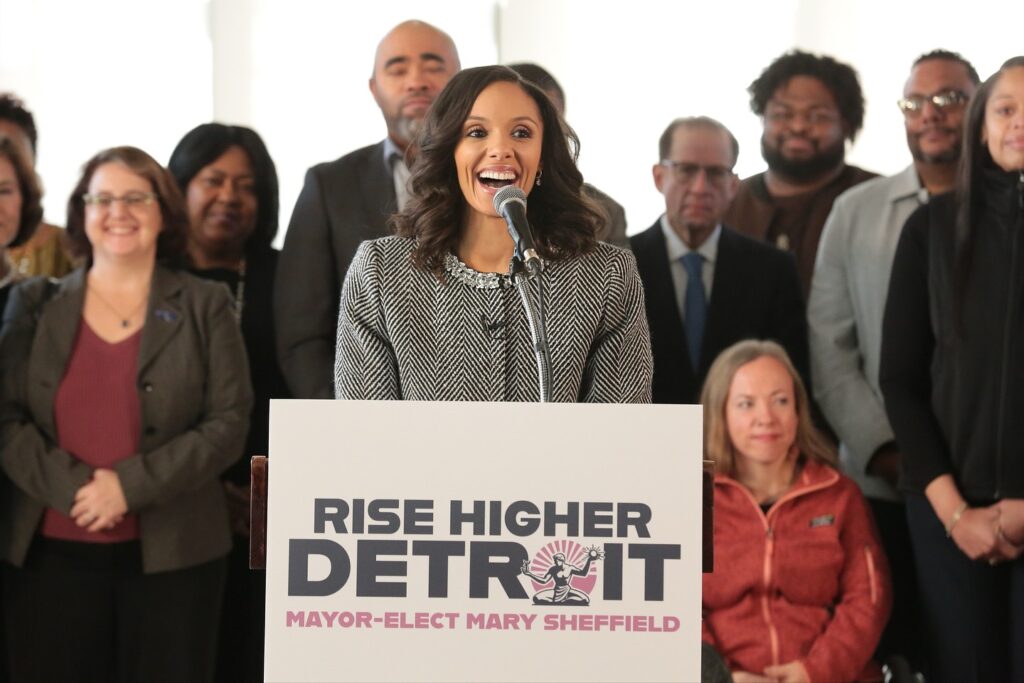 Mayor-elect Sheffield speaking at a podium for the Rise Higher Detroit transition team. Behind her is various team members, with Megan standing at left.