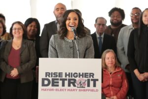 Mayor-elect Sheffield speaking at a podium for the Rise Higher Detroit transition team. Behind her is various team members, with Megan standing at left.