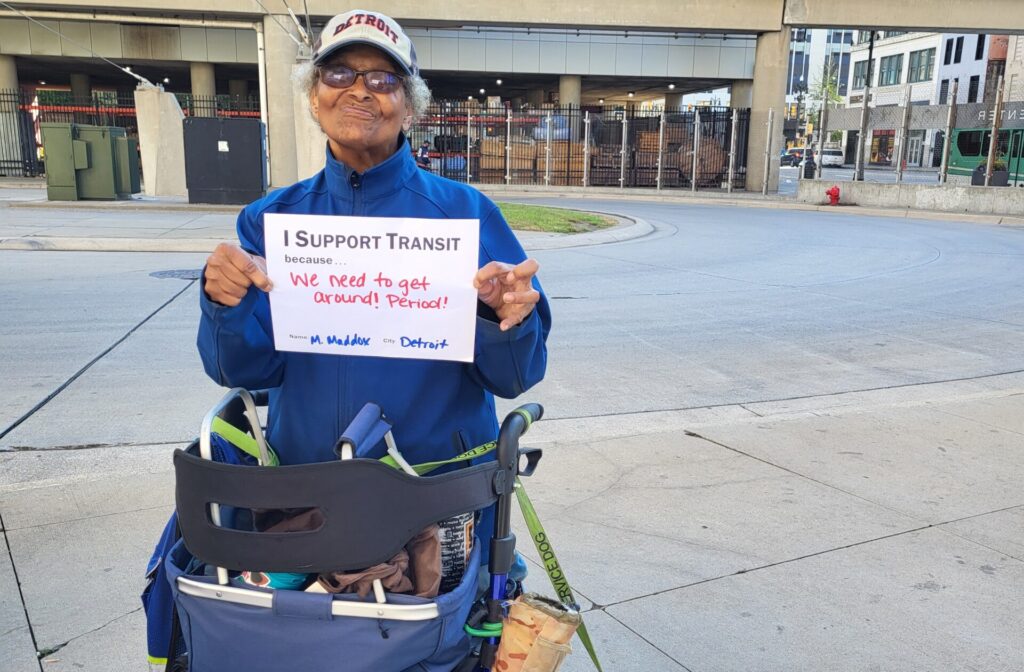 Magurite Maddox at Rosa Parks Transit Center, holding an i support transit sign with her service dog