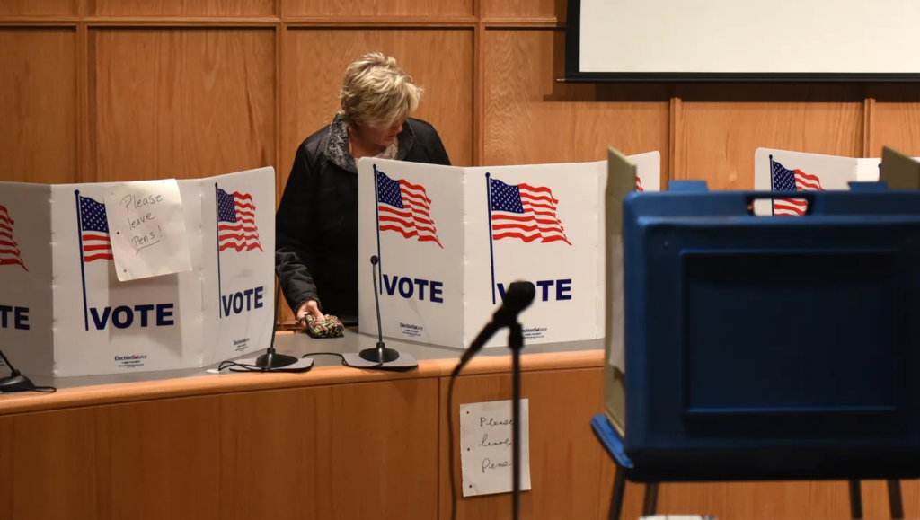 Voter voting behind a trifold, with the American flag on it and "VOTE" in text below