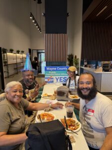 A group of african-american people sitting at a table, with a yard sign in the background saying "Wayne County public transportation millage, vote yes!"
