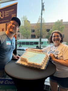 Two white people holding up a cake with Wayne County on it. A DDOT bus in the background
